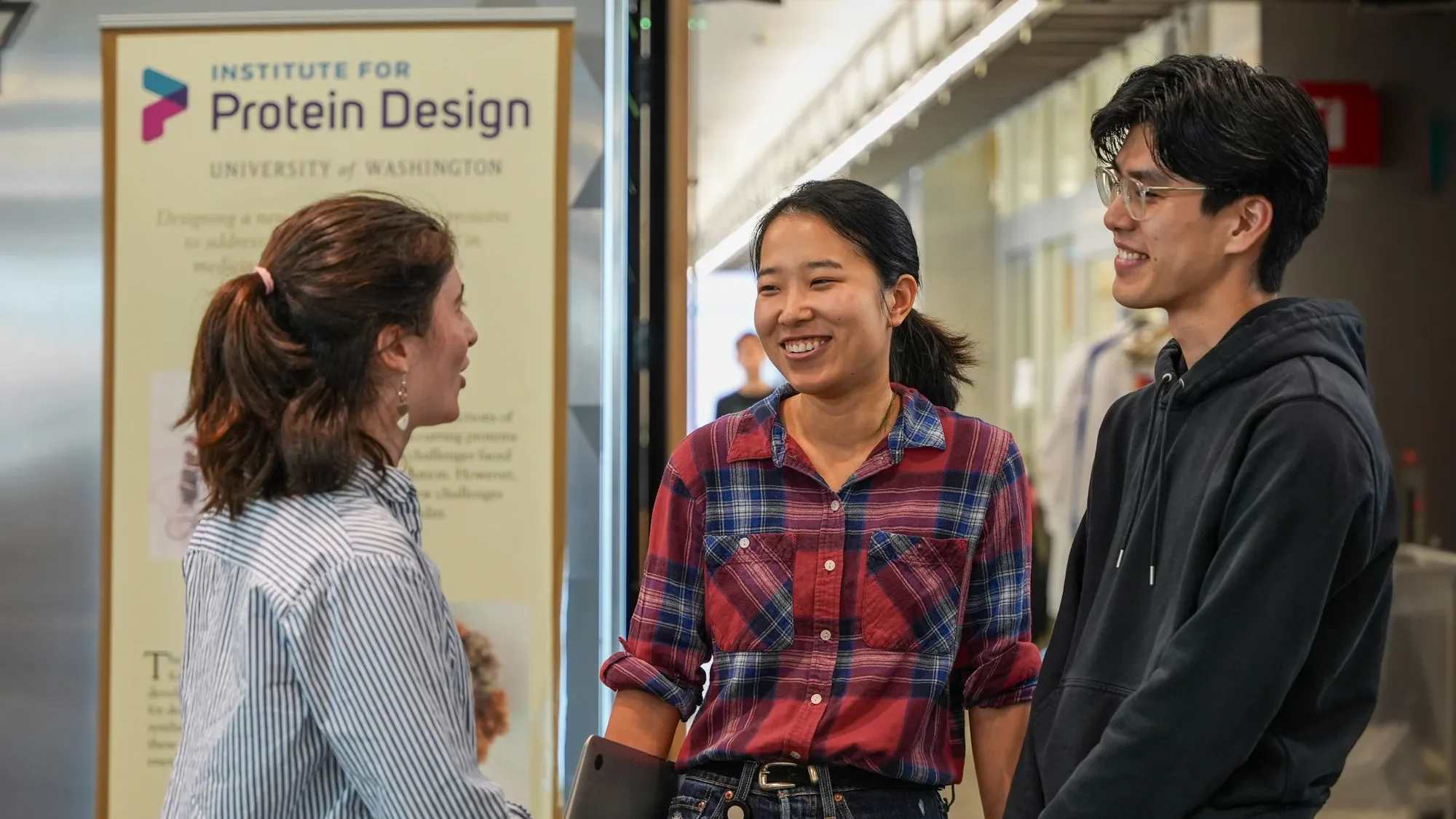 Three Institute for Protein Design researchers talking and smiling in a hallway near the IPD banner.